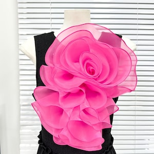 May include: A black sleeveless top featuring a large, layered, hot pink organza flower appliqué on the front. The flower is constructed from multiple translucent, ruffled petals that create a voluminous, dimensional floral design.
