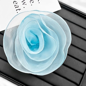 May include: A light blue fabric flower accessory crafted from sheer, translucent organza material. The flower features multiple layered, ruffled petals arranged in a circular, rose-like pattern with finished, stitched edges throughout the design.