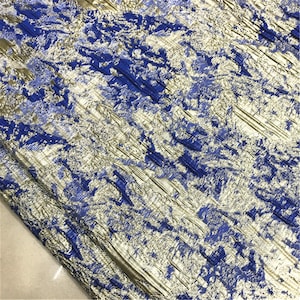 May include: Close-up of a textured fabric with a metallic sheen. The fabric features a pattern of blue and gold, with a crinkled appearance. The design has a layered effect, creating depth and visual interest. The fabric is likely used for apparel or home decor.