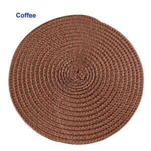 May include: A round, woven placemat in a warm brown hue. The placemat is made of tightly woven fibers, creating a textured surface. The word "Coffee" is printed in blue in the upper left corner. This placemat is suitable for dining and kitchen decor.