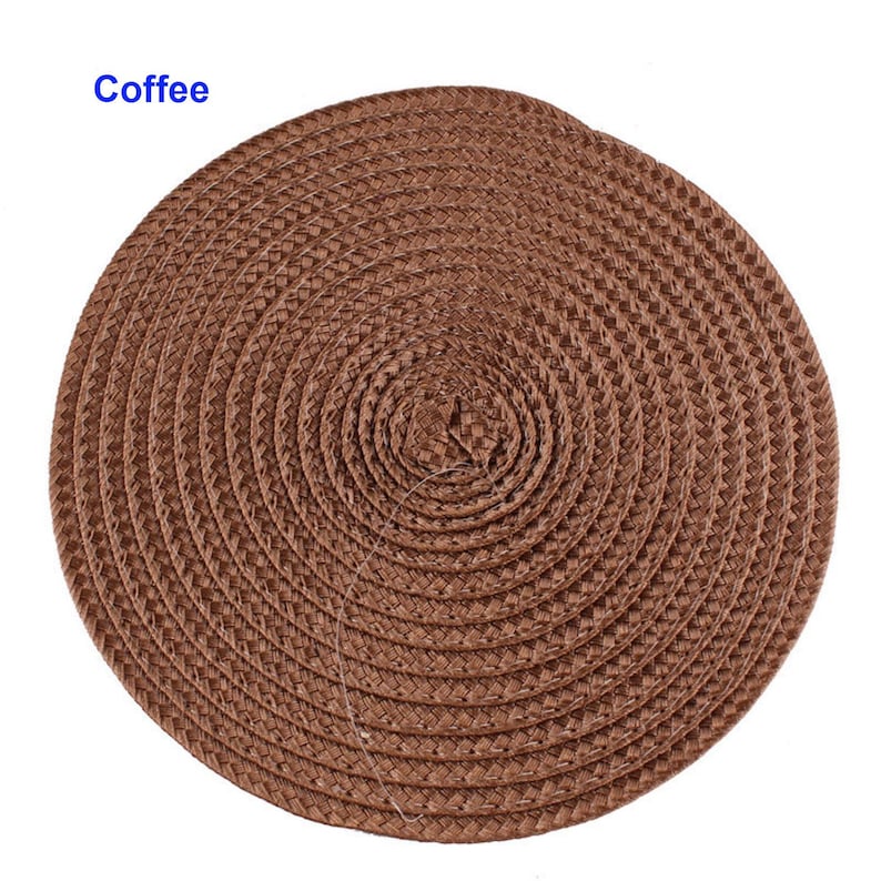 May include: A round, woven placemat in a warm brown hue. The placemat is made of tightly woven fibers, creating a textured surface. The word "Coffee" is printed in blue in the upper left corner. This placemat is suitable for dining and kitchen decor.
