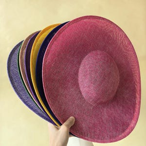 May include: A collection of wide-brimmed hats in various colors, including pink, purple, yellow, and blue. The hats have a textured appearance and a rounded crown. The hats are held by a hand, showcasing their size and shape.