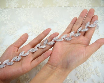 May include: A silver rhinestone trim with a leaf design. The trim is made of small, clear rhinestones that are set in a silver metal base. The trim is flexible and can be used to decorate a variety of items, such as clothing, shoes, bags, and home decor.