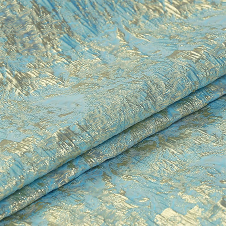 May include: Close-up of a folded fabric with a textured pattern. The fabric has a light blue base color with gold metallic accents. The metallic elements create a shimmering effect, adding depth and visual interest to the textile.