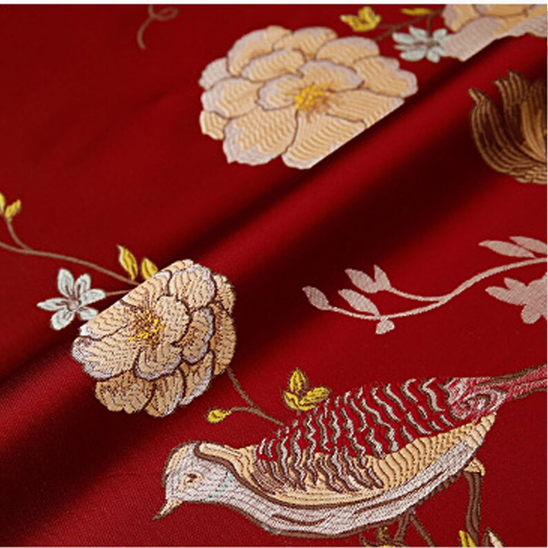 May include: Red fabric with an embroidered design of a bird and flowers. The bird is brown and white with red accents. The flowers are white with yellow centers.