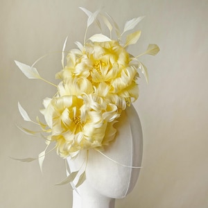 May include: A pale yellow floral headpiece with delicate white feathers and accents. The headpiece features two large, ruffled flower-like designs with small yellow details in the centre. The accessory is displayed on a white mannequin head.