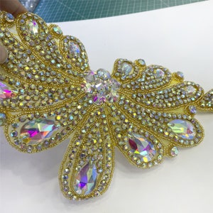 May include: A large, gold-colored, butterfly-shaped embellishment with iridescent rhinestones. The embellishment is made of small, round beads and larger, teardrop-shaped rhinestones.