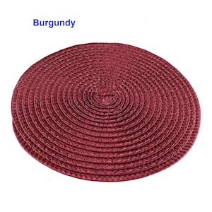 May include: A round burgundy placemat with a woven texture. The placemat is circular, with a spiral design radiating from the center. The word "Burgundy" is printed in blue in the upper left corner.