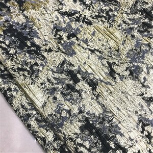 May include: Close-up of a textured fabric with a pattern of black, gray, and gold metallic accents. The fabric has a crinkled appearance with diagonal lines and a subtle sheen, suitable for apparel or home decor.