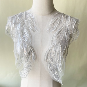 May include: A pair of white beaded and sequined applique embellishments in the shape of feathers. The embellishments are on a sheer mesh fabric.