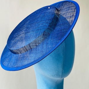 May include: A vibrant blue hat with a wide brim and a slightly angled crown. The hat is made of a woven material, with a darker blue trim around the edge. A small, white decorative element is visible on the crown. The hat is displayed on a blue mannequin head.