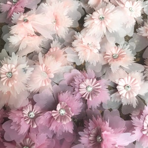 May include: Close-up of a pink and white fabric with 3D flower embellishments. The flowers are made of layers of sheer fabric and have a rhinestone center.