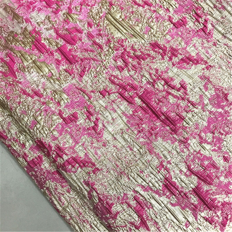 May include: Close-up of a textured fabric with a metallic gold and hot pink pattern. The fabric has a crinkled appearance with diagonal lines and irregular shapes. The colors create a striking contrast, suitable for apparel or home decor.