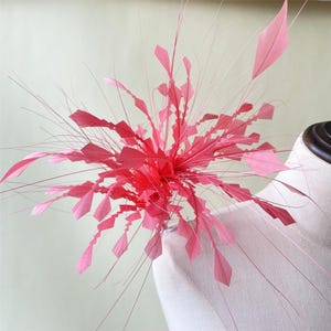 Könnte beinhalten: Ein leuchtend pinkfarbener Fascinator, der eine Explosion aus geschichteten, geometrisch geformten Blütenblättern und zarten, fedrigen Strähnen zeigt. Das Kopfstück wird auf einer Schaufensterpuppe präsentiert.