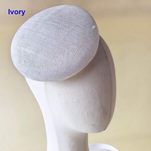 May include: Ivory-colored, oval-shaped hat with a textured surface. The hat is attached to a mannequin head. The word "Ivory" is printed in blue in the upper left corner. A small, white pearl is attached to the hat.