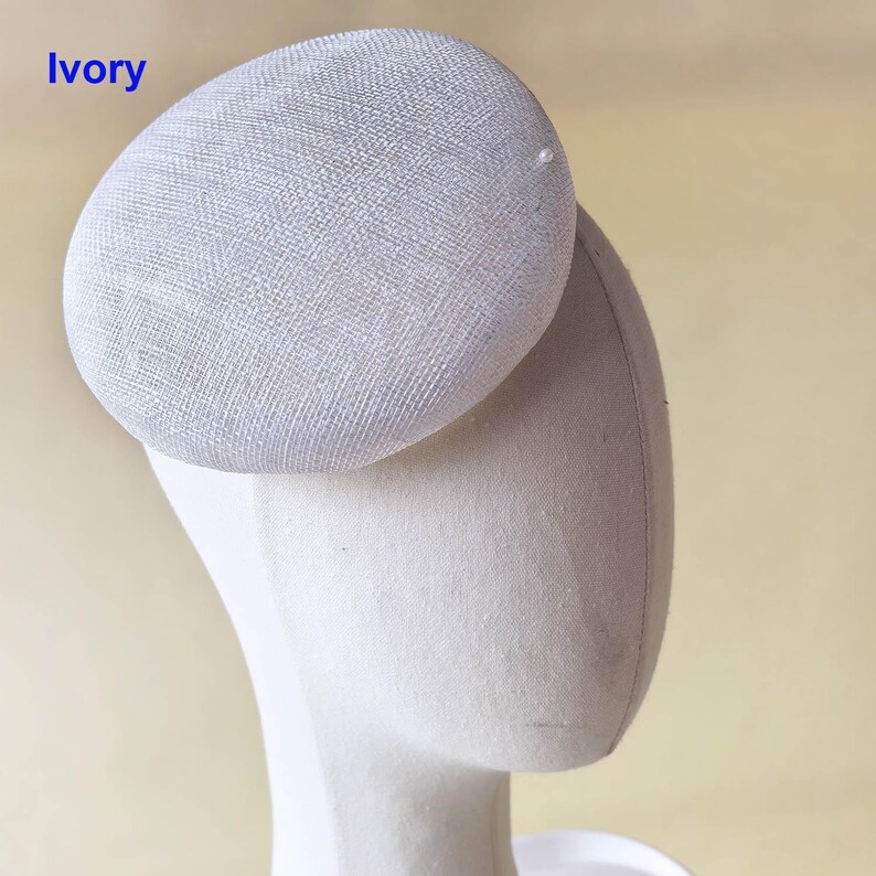 May include: Ivory-colored, oval-shaped hat with a textured surface. The hat is attached to a mannequin head. The word "Ivory" is printed in blue in the upper left corner. A small, white pearl is attached to the hat.