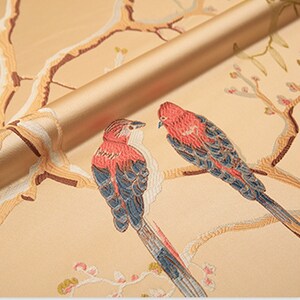May include: A close-up of a beige fabric with an embroidered design of two birds perched on branches. The birds have red and blue plumage, and the branches are adorned with delicate white flowers.