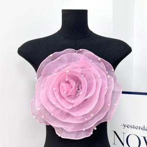 Peut inclure: Une grande fleur en organza rose à plusieurs couches, dotée de pétales translucides aux bords volantés. La fleur est ornée de petites perles blanches rondes réparties sur la surface des pétales, créant un motif floral texturé et détaillé.