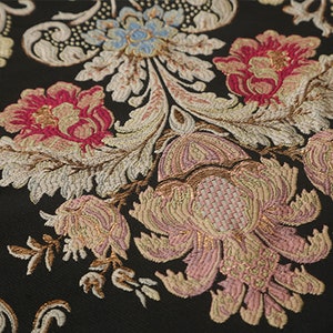 May include: Close-up of a black fabric with a floral pattern. The fabric features pink, white, and gold flowers with intricate details. The background is black.