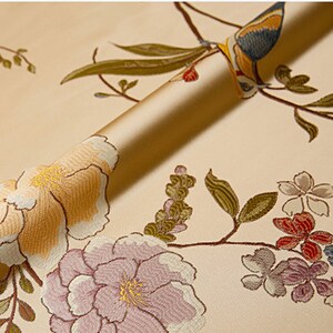 May include: A close-up of a beige fabric with an embroidered floral pattern. The fabric features pink, yellow, and green flowers, as well as a blue and yellow bird.