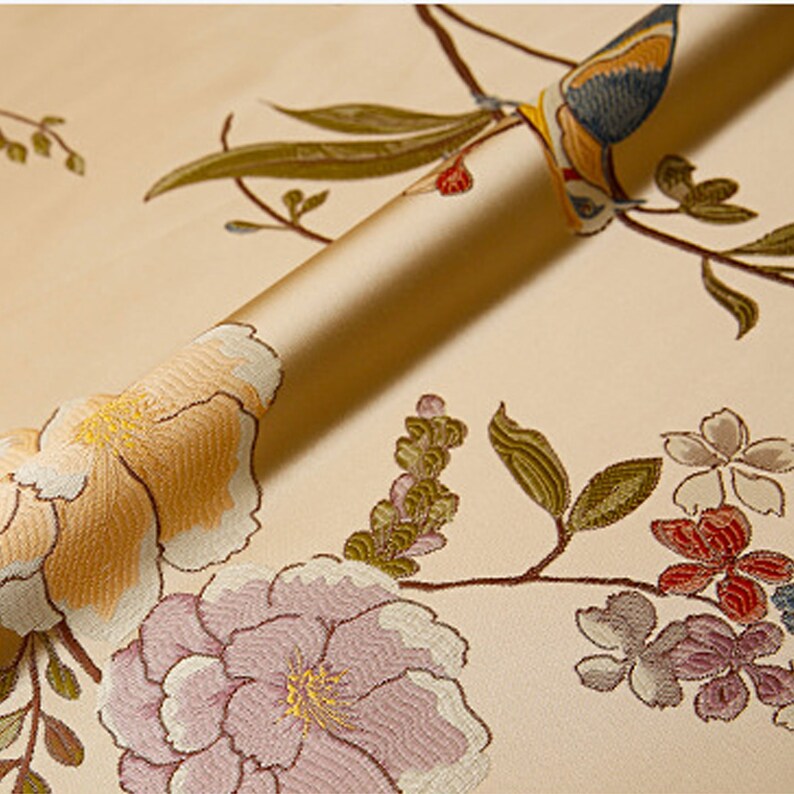 May include: A close-up of a beige fabric with an embroidered floral pattern. The fabric features pink, yellow, and green flowers, as well as a blue and yellow bird.