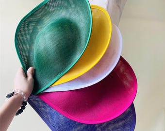 May include: A collection of colorful, wide-brimmed hats in shades of green, yellow, white, pink, and blue. Each hat has a mesh-like texture and a contrasting trim. The hats are held in a hand, showcasing their variety.
