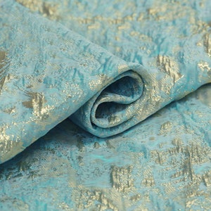 May include: Close-up of a folded fabric with a textured surface. The fabric is a light teal color with a gold metallic pattern. The gold pattern has a floral or abstract design. The fabric is likely used for apparel or home decor.