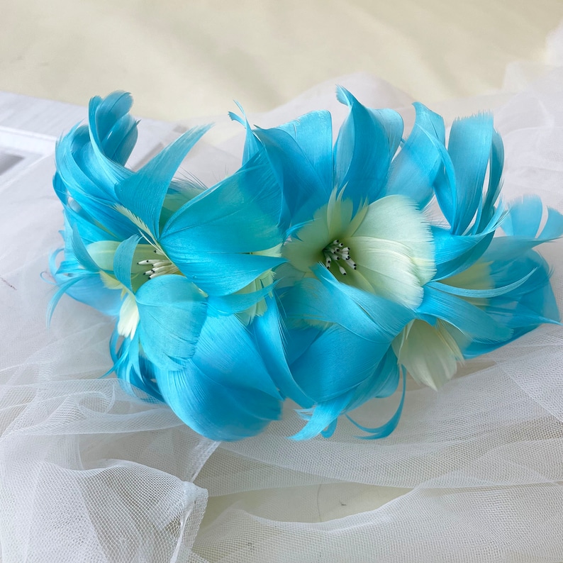 May include: A blue feather flower headband with white accents. The headband is made of feathers and is perfect for adding a touch of whimsy to any outfit.