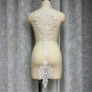 May include: White lace applique with a floral design. The applique is shaped like a teardrop and has a delicate, intricate pattern. It is perfect for adding a touch of elegance to any garment.