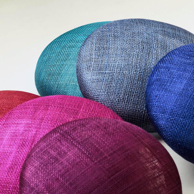 May include: A collection of colorful, round, fabric-covered hat bases in various shades. The hats are in shades of teal, blue, purple, and red. The fabric has a woven texture, and the hats are arranged in a semi-circle.