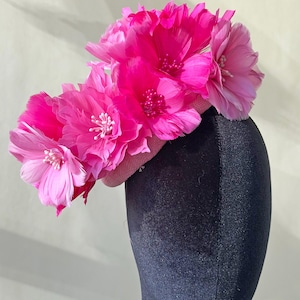 May include: A striking pink fascinator with large, feathery flower decorations. The flowers are a vivid fuchsia, with intricate detailing. The base of the fascinator is a muted pink, creating a bold contrast. Ideal for events and special occasions.