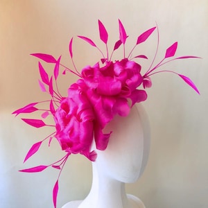 Può includere: Un copricapo fucsia vibrante con due grandi fiori di tessuto arricciato e delicate foglie appuntite. Il copricapo è esposto su una testa di manichino bianca, evidenziandone il design. Un accessorio per occasioni speciali.