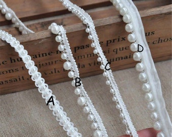 May include: Four different styles of white pearl trim are displayed. The trims vary in design, with some featuring dangling pearls and others with pearls sewn directly onto the trim. The trims are ideal for embellishing clothing or craft projects.