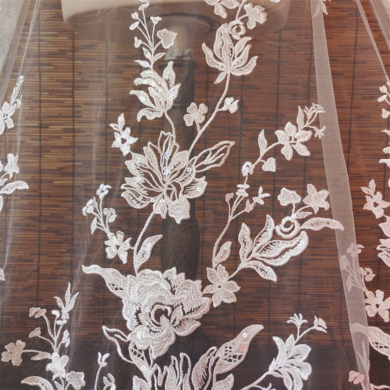 May include: White floral lace fabric with sequins. The lace features a delicate floral pattern with small sequins scattered throughout. The fabric is sheer and lightweight.