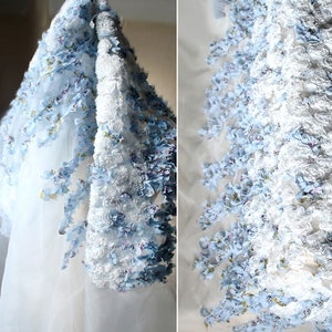 May include: White sheer fabric with a blue and white floral trim. The trim is made of 3D flowers and has a ruffled texture.