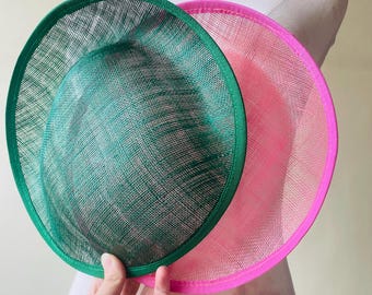 May include: Two circular hats, one emerald green and one bright pink, are displayed. The hats have a woven texture and a contrasting trim. The green hat is in front, partially obscuring the pink hat. The hats are on a mannequin.