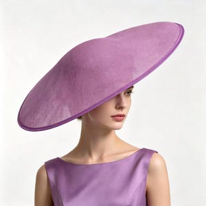 Peut inclure: Un chapeau large de couleur lavande avec une calotte arrondie et lisse et un bord structuré légèrement asymétrique. Le chapeau présente une texture finement tissée et un liseré lavande assorti sur le bord, porté avec une robe sans manches.