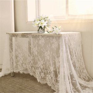 Peut inclure: Nappe en dentelle blanche avec un motif floral délicat. La nappe est drapé sur une table et a un bord festonné.