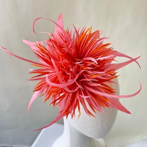 Puede incluir: Un fascinador de plumas de color coral vibrante con detalles en naranja. El tocado presenta un grupo central denso y puntiagudo rodeado de delicadas plumas curvas en tonos rosa y coral. El accesorio está montado sobre una cabeza de maniquí blanca.
