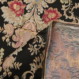 May include: Close-up of black fabric with a gold and pink floral pattern. The fabric has a textured, woven look and feel. The fabric is folded over to show the back side, which is a plain, woven fabric.