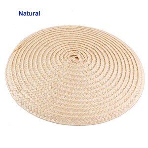 May include: A round, woven placemat in a natural beige color. The placemat is made of tightly woven straw or similar material, with a spiral pattern radiating from the center. The word "Natural" is printed in blue in the upper left corner.