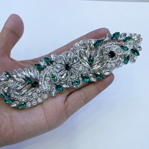 Puede incluir: Una aplicación de cuentas verdes y blancas con diamantes de imitación transparentes y verde oscuro. La aplicación está sobre una base de tela transparente.
