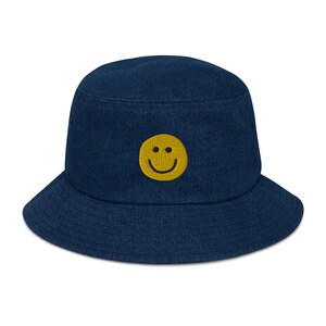 Può includere: Un cappello a secchiello blu navy con una faccina sorridente gialla ricamata sul davanti.