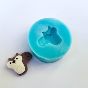 May include: A light blue silicone mold with a squirrel design next to a finished squirrel-shaped embellishment. The squirrel is brown, white, and beige with a small heart design. The mold is round.