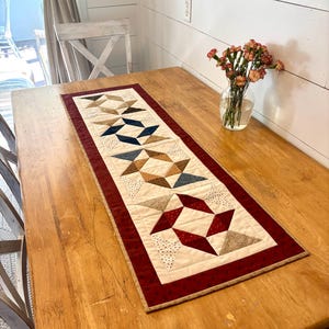 May include: A rectangular quilted table runner featuring a series of four star-patterned blocks in shades of cream, navy blue, tan, and deep red. The runner is framed by a wide, dark red border with a subtle pattern and a thin tan outer edge.