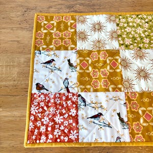 May include: A quilted square featuring a patchwork design with various patterns. The patterns include floral, sunburst, and bird motifs in shades of gold, white, orange, and green. The edges are finished with a yellow border.