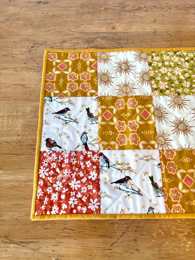 May include: A quilted square featuring a patchwork design with various patterns. The patterns include floral, sunburst, and bird motifs in shades of gold, white, orange, and green. The edges are finished with a yellow border.