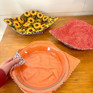 May include: Three fabric bowl cozies in different patterns. One features a sunflower print, another has a red and white floral pattern, and the third is a solid orange with a white floral trim. The cozies are designed to protect surfaces from hot dishes.