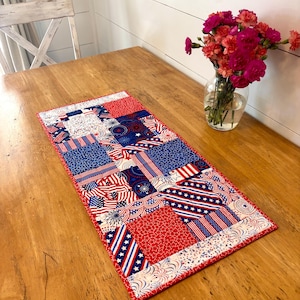 May include: A rectangular table runner with a patchwork design in red, white, and blue. The runner features patterns of American flags, stars, and stripes. It is placed on a wooden table, with a vase of pink flowers in the background.