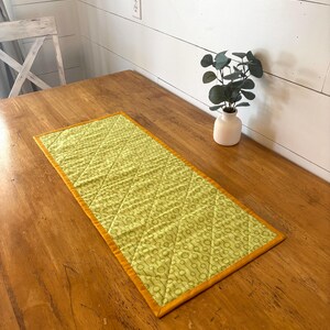 May include: A rectangular, quilted table runner with a light green floral pattern and a gold border. The runner is placed on a wooden table. A small white vase with greenery is in the background.